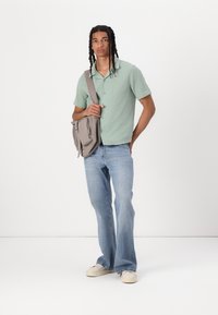 Lichtgroen kortebloesje met knopen, gecombineerd met vervaagde blauwe flared jeans en een beige crossbody tas. Eenvoudige textuur en een relaxte pasvorm.