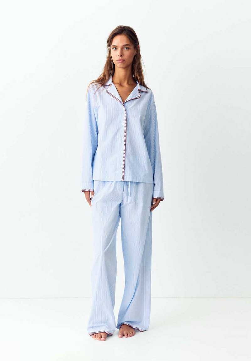 Femme debout, pieds nus, portant un ensemble pyjama rayé bleu clair à manches longues avec des coutures foncées sur le col, les poignets et les ourlets, sur fond blanc.