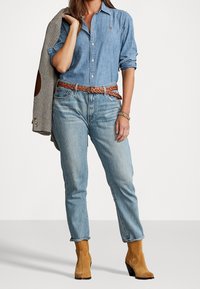 Ljusblå denimskjorta, ljusblå jeans, brun flätad läderbälte, bruna mockastövlar, herringbone-blazer draperad över axeln.