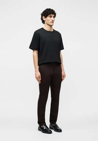 Jeune homme debout, portant un t-shirt noir uni à manches courtes, un pantalon slim foncé et des mocassins en cuir noir sur fond blanc.