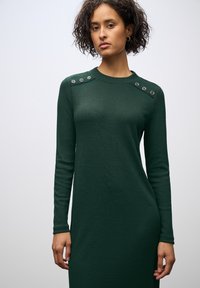 Abito a maniche lunghe, di un verde scuro a costine con bottoni a pressione sulle spalle. Presenta un classico scollo tondo e una silhouette aderente.