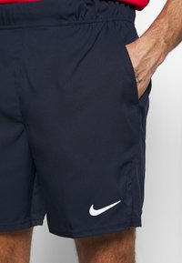 Pantalones cortos marinos hechos de tela ligera, con una cinturilla elástica y un bolsillo lateral. Un logo blanco de Nike se muestra en el dobladillo.