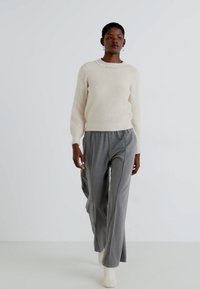 Beige gestrickter Pullover mit Rippstruktur, kombiniert mit lockeren grauen Hosen. Das Model trägt cremefarbene Stiefeletten; einfaches Design mit minimalen Akzenten.