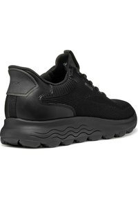 Svart sneaker med en texturerad tygöverdel och släta läderdetaljer. Tjock, vadderad sula med en vågmönster på undersidan.