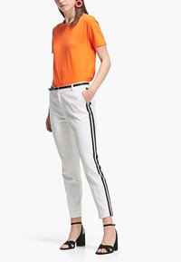 Pomarańczowa koszulka z krótkim rękawem połączona z białymi spodniami na miarę z czarnymi paskami po bokach oraz czarnymi sandałami na obcasie.