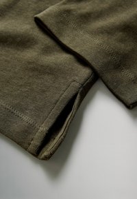 Indumento in cotone verde oliva con polsini a coste e una trama liscia, caratterizzato da cuciture precise e da un leggero spacco all'orlo.