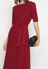 Ein zerdrücktes rotes Midi-Kleid mit kurzen Ärmeln, das eine gebundene Taille und einen strukturierten Stoff aufweist. Ergänzt durch eine schwarze, gefaltete Clutch.