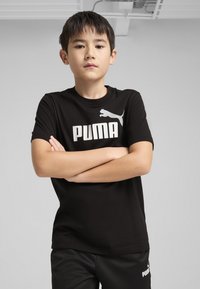 T-shirt noir à manches courtes avec grand imprimé blanc "PUMA" et logo. Texture lisse, design classique à col rond, associé à un pantalon noir.