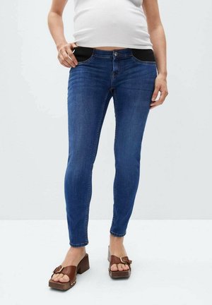 Kvinde iført blå skinny jeans med justerbare sorte elastiske taljeindlæg og brune platformssandaler, stående mod en ensfarvet baggrund.