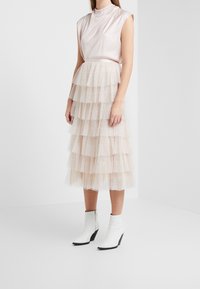 Haut en satin rose clair avec manches courtes, associé à une jupe beige en tulle à plusieurs couches, longueur genou, ornée de petits pois et d'une ceinture. Bottines blanches à la cheville.