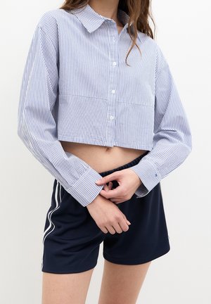 Woman wearing a cropped, long-sleeve, blue-and-white striped button-up shirt and navy shorts with white side stripes, hands clasped in front.
