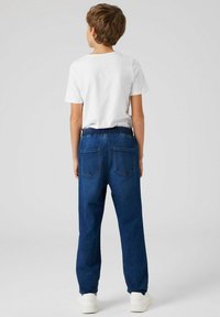 Mėlynos džinsinės treniruotės kelnės su elastinga juosmens juosta, dviem galinėmis kišenėmis ir laisvu siluetu, derinamos su paprastu baltu marškinėliu ir baltais sportbačiais.