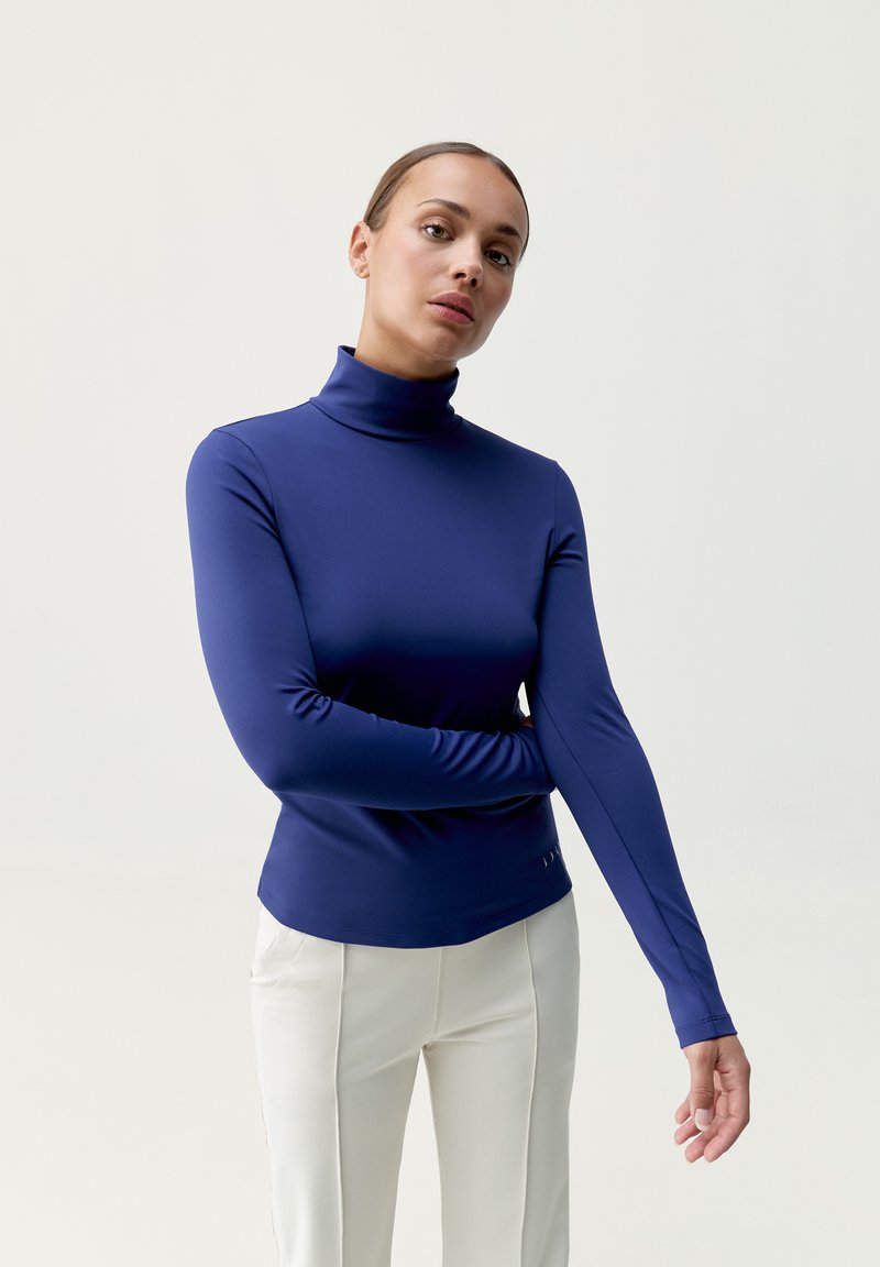 Marinblå långärmad polotröja iStretchy material, med en kroppsnära design och slät yta, kombinerad med ljusa byxor.