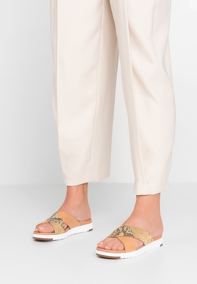 Pieds chaussés de sandales slides beige à motif serpent avec semelles blanches, assorties à un pantalon court couleur crème.