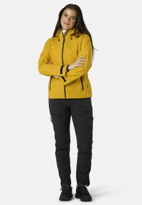 Giacca impermeabile gialla con tasche a zip e dettagli neri, abbinata a pantaloni neri con tasche cargo. Scarpe robuste completano l'outfit.