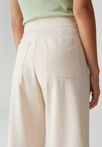 Beige wijde broek met een soepele textuur, voorzien van twee achterzakken en een hoge tailleband, weergegeven vanuit de achterkant.