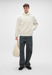 Krema flis dukserica s utisnutim logotipom "BOSS", uparen s tamnim trapericama i mekanom torbom krem boje. Crne slip-on cipele.