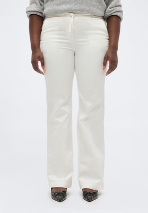 Persoană care poartă pantaloni de culoare albă-crem, cu talie înaltă și croială dreaptă, având șnur în talie și încălțări negre cu vârf ascuțit, stând pe un fundal alb.