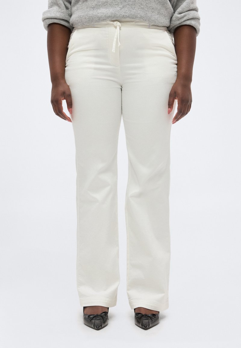 Άτομο που φορά εκρού παντελόνι με ίσια γραμμή, ψηλή μέση και κορδόνι στη μέση, και μαύρα παπούτσια με μυτερή μύτη, στέκεται μπροστά σε λευκό φόντο.