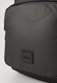Sac à dos en tissu noir avec une texture lisse, arborant un patch logo rectangulaire et une poche zippée à l'avant. Design minimaliste.