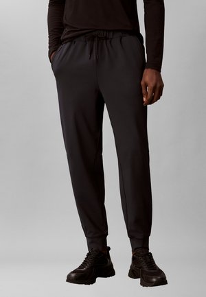 Homme portant un pantalon de jogging noir avec taille à cordon et des baskets noires, debout devant un fond gris uni.