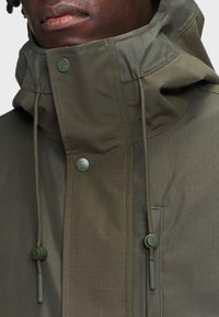 Gros plan sur une personne à la peau foncée portant une veste à capuche vert olive avec rabat à boutons et cordons ajustables.