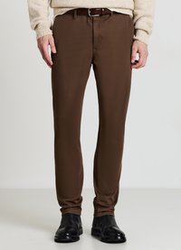 Braune Hose mit glatter Textur, schmaler Passform und Gürtelschlaufen. Kombiniert mit einem beigen Pullover und schwarzen Lederschuhen.