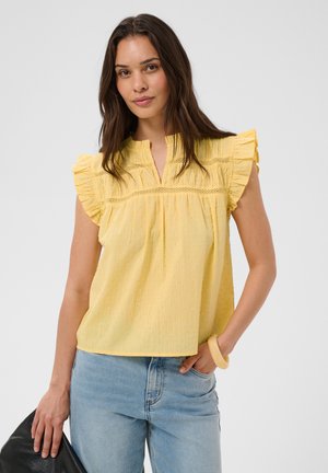 Femme portant un chemisier sans manches jaune à volants et un jean bleu clair, tenant un sac noir dans une main et l'autre main dans la poche.