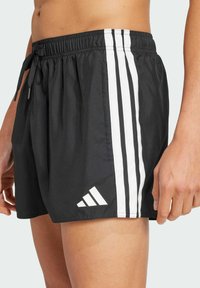 Crne kupaće gaće s podesivim strukom, s tri vertikalne bijele pruge duž bokova i bijelim Adidas logotipom na donjem rubu.