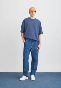 Přetahovaný námořnický modrý tričko, světle modré džíny s viditelnými kapsami, bílé tenisky a oranžová baseballová čepice. Jednoduchý design, volný střih.