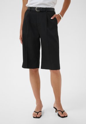 Frau trägt schwarze, knielange, maßgeschneiderte Shorts mit Gürtel und schwarze Sandalen mit Absatz, steht mit einer Hand in der Tasche.