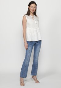 Top bianco senza maniche con inserti in pizzo, dettagli plissettati e scollatura a chiave, abbinato a jeans blu aderenti e sandali con tacco trasparente.