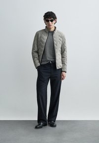 Vaaleanharmaa tikattu takki pystykauluksella, päällä teksturoitu tummanharmaa paita ja mustat housut. Mustat aurinkolasit ja kengät viimeistelevät asun.