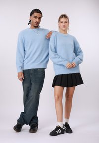 Svetlo modri, puhasti puloverji iz pletenine z nizkimi rameni. Model na levi v temnih kavbojkah in črnih čevljih, model na desni v črni plisirani krilu in supergah.