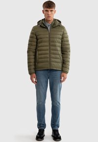 Letthældig olivengrøn pufferjakke med hætte, der har en kviltet tekstur og frontlynlås. Bærer den med blå jeans og sorte sneakers.