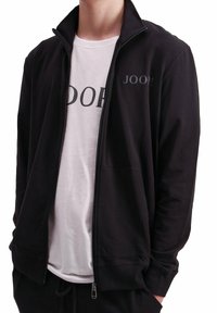 Schwarze Zip-Jacke mit Stehkragen und seitlichen Taschen. Trägt ein weißes T-Shirt mit dem "JOOP!"-Logo in dunklen Buchstaben.