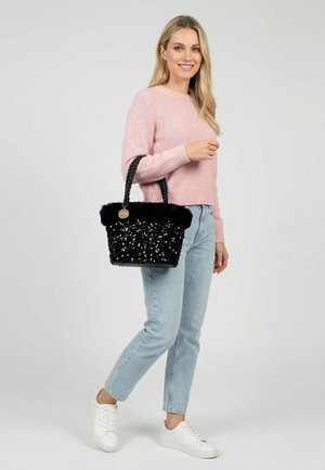 Mujer con suéter rosa y vaqueros azul claro sostiene un bolso negro con lentejuelas y asas trenzadas, lleva zapatillas blancas y está de pie sobre un fondo gris.