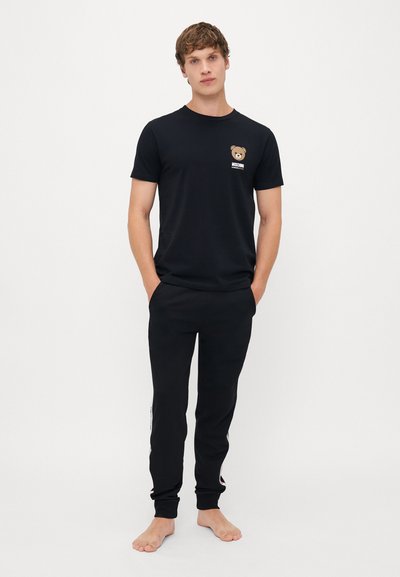 Jovem a pé descalço com as mãos nos bolsos, vestindo uma t-shirt preta da Moschino com o logótipo do urso de pelúcia e calças pretas a condizer.