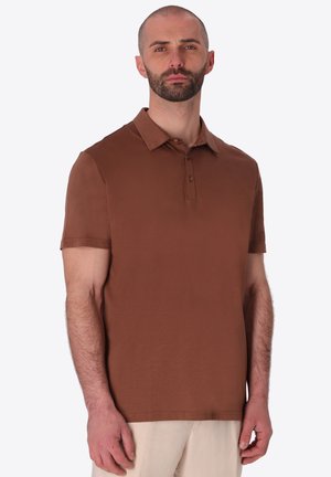 Uomo con la barba che indossa una polo marrone a maniche corte e pantaloni beige chiaro, in piedi davanti a uno sfondo chiaro e semplice.