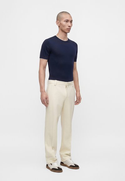 Homme debout portant une chemise ajustée bleu marine à manches courtes, un pantalon couleur crème et des baskets blanches avec des touches noires et marron.