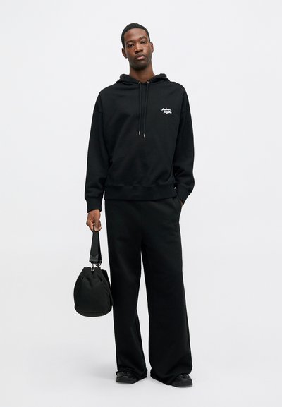 Hanorac negru cu logo pe față, buzunar tip cangur și șnur de ajustare. Asortat cu pantaloni negri largi și o geantă neagră. Textură fină.