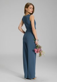Teal jumpsuit met een aansluitend lijfje en wijde broekspijpen. Glad materiaal, mouwloos ontwerp en een subtiele naad aan de achterkant. Houdt een boeket bloemen vast.