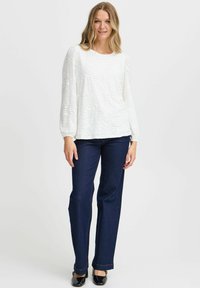 Vit långärmad topp med texturerad vågmönster, matchad med mörkblå raka jeans och svarta skor. Enkel, avslappnad outfit.