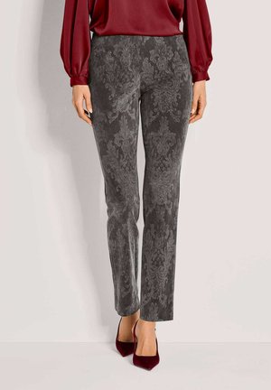 Vrouw in grijze, met een patroon bedrukte fluwelen broek, rode blouse met lange mouwen en rode puntige pumps, staande tegen een lichte achtergrond.