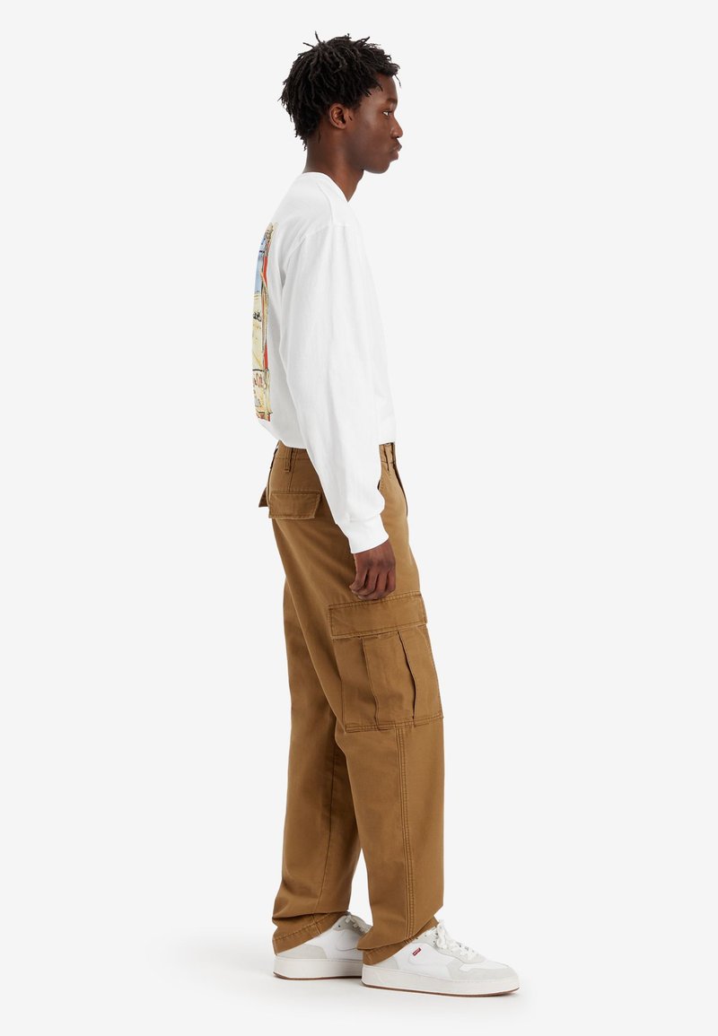 Camisa blanca de manga larga con un gráfico en la parte posterior, combinada con pantalones cargo beige que tienen bolsillos laterales, usados con zapatillas blancas.