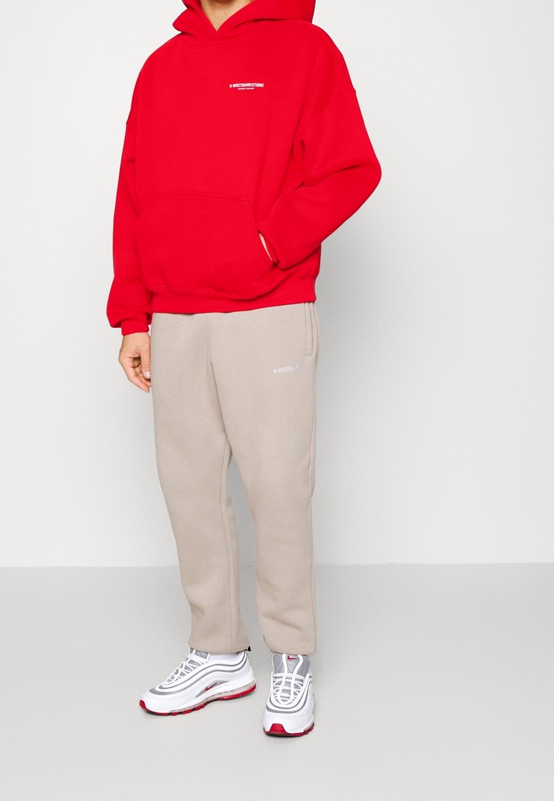 Personne portant un sweat à capuche rouge avec une poche frontale, un pantalon de jogging beige et des baskets blanches avec des touches rouges et grises, debout devant un fond uni.