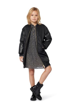 Schwarze Bomberjacke mit goldener Reißverschluss, getragen über einem gefalteten Kleid mit schwarz-weißer Musterung, kombiniert mit glänzenden schwarzen Stiefeletten.