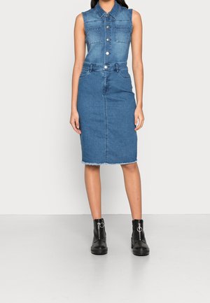Robe en jean bleu sans manches avec boutons devant, poches poitrine, ourlet effiloché à hauteur des genoux, portée avec des bottines noires à fermetures éclair argentées rondes.