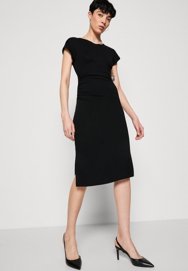Czarna sukienka do kolan z krótkimi rękawami, dopasowany krój, subtelne marszczenie w talii i boczne rozcięcie. Noszona z czarnymi szpilkami w czubek.
