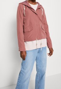Hellrosa Kapuzenjacke mit Kordelzug und zwei Vorderentaschen, darüber ein pinker Pullover, kombiniert mit hellblauen Jeans mit weitem Bein.